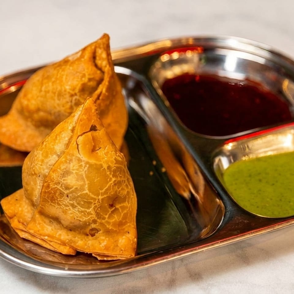 Two golden samosas served in a stainless steel tray with green chutney and dark red dipping sauce.