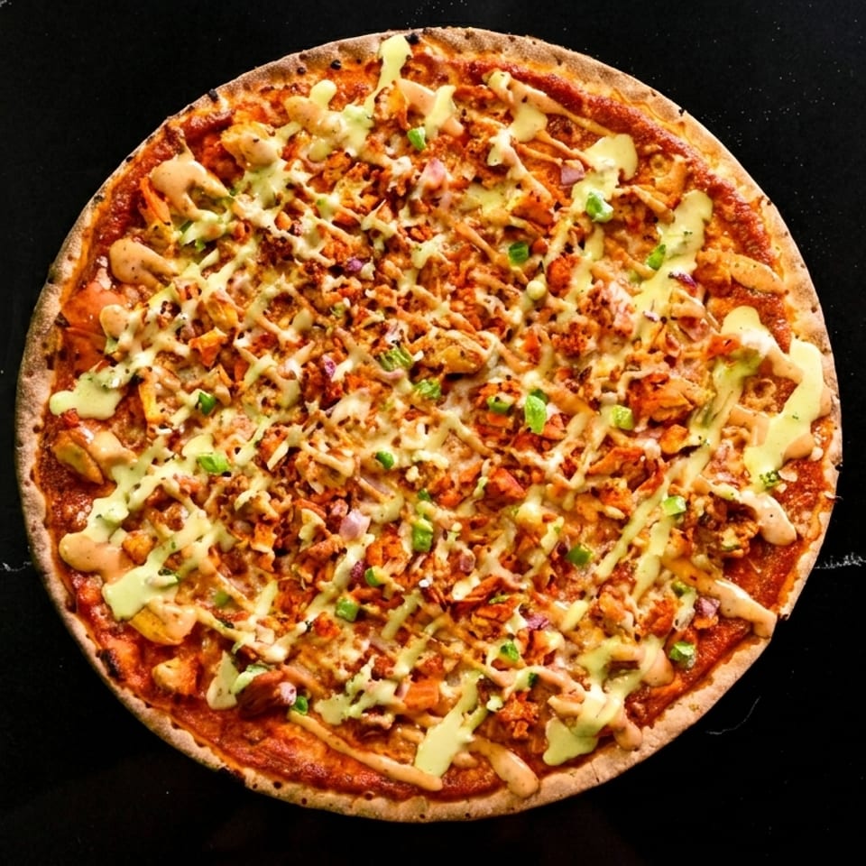 Pizza topped with chicken, vegetables, melted cheese, and drizzled sauces on a black marble surface.